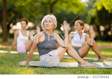Senior woman performing Matsyendrasana during group yoga practice outdoors 121738182