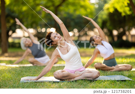 Portrait of caucasian woman sitting on sports mat and making yoga meditation in lotus pose and hands up with group together in park at daytime 121738201