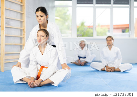 Young woman in kimono stretching in butterfly pose with assistance of trainer 121738301