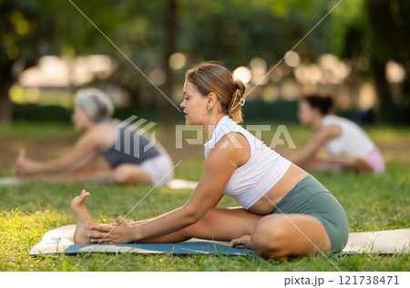 Portrait of woman sitting on sports mat and making yoga exercises with group together in green park at daytime 121738471