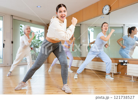 Group of happy young mature sportive women exercising dancing moves with instructor in modern gym studio Group of happy young mature sportive women exercising dancing moves with instructor in modern gym studio 121738527