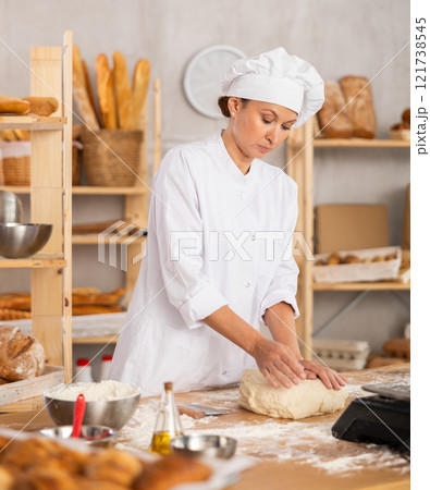 Experienced female baker preparing raw dough on kitchen table in bakery 121738545