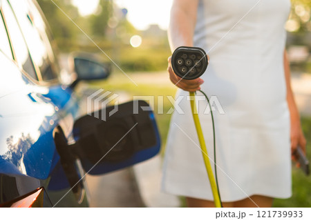 Woman holding charger plug in front of her to charge the electric car in the street Woman holding charger plug in front of her to charge the electric car in the street 121739933