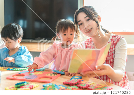 保育園・幼稚園・託児所・保育所で園児の女の子に絵本を読み聞かせする女性保育士・保母さん・シッター 保育園・幼稚園・託児所・保育所で園児の女の子に絵本を読み聞かせする女性保育士・保母さん・シッター 121740286