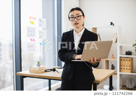 Portrait of young Asian businesswoman standing with laptop in office environment. She wears black suit and glasses, conveying professionalism and confidence. Bright modern workspace with large windows Portrait of young Asian businesswoman standing with laptop in office environment. She wears black suit and glasses, conveying professionalism and confidence. Bright modern workspace with large windows 121742266
