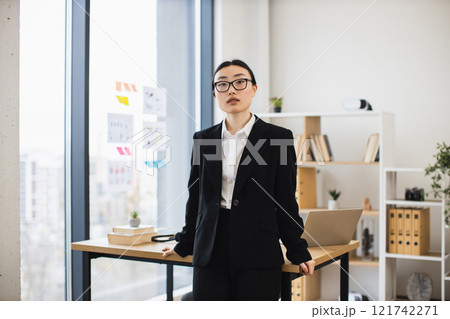 Portrait of confident young Asian businesswoman in office wearing glasses and formal suit, standing by desk. Professional female projecting confidence and leadership. Portrait of confident young Asian businesswoman in office wearing glasses and formal suit, standing by desk. Professional female projecting confidence and leadership. 121742271