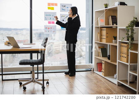 Asian businesswoman in professional attire multitasking while talking on phone and writing on glass wall in bright office setting. 121742280