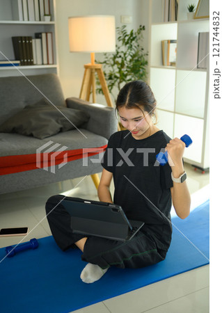 Young asian woman lifting a light dumbbell and using digital tablet on yoga mat 121744832