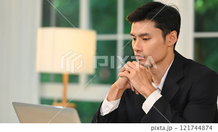 Thoughtful millennial entrepreneur with his hands clasped under his chin working on laptop 121744874
