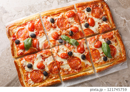 Savory open pie made of puff pastry with pepperoni sausage, olives, mozzarella and tomato sauce close-up on parchment. horizontal top view 121745759