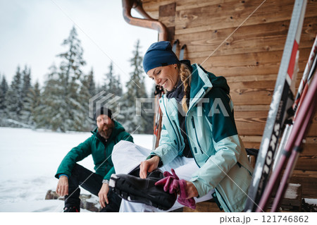 Couple preparing for cross country skiing in winter nature, putting boots on. Sitting in front cabin. 121746862