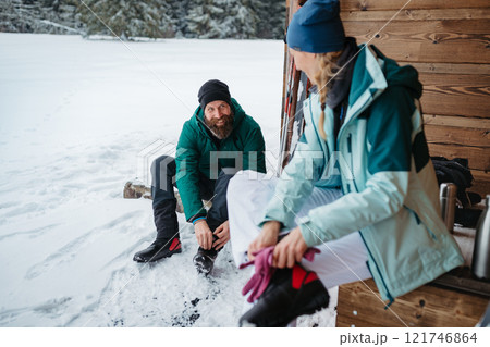 Couple preparing for cross country skiing in winter nature, putting boots on. Sitting in front cabin. Couple preparing for cross country skiing in winter nature, putting boots on. Sitting in front cabin. 121746864