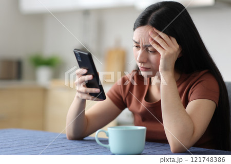 Worried asian woman checking smart phone in the kitchen Worried asian woman checking smart phone in the kitchen 121748386