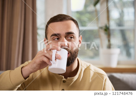 Healthy young man drinking hot mineralizing, vitaminizing drink sitting on sofa in cozy living room at home. Healthy young man drinking hot mineralizing, vitaminizing drink sitting on sofa in cozy living room at home. 121748476