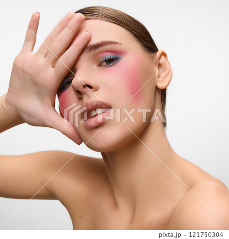 Close-up of model with vibrant pink blush and pastel eye makeup, gracefully covering part of her face with hand against white studio background. Concept of natural beauty. 121750304