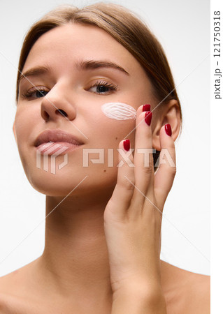 Smiling attractive woman applying cream to her cheek, symbolizing moisturizing and skincare routines against white studio background. Concept of natural beauty. 121750318