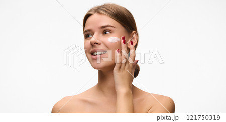 Horizontal image of beautiful woman spreading cream on her cheek, focusing on hydration and skin health against white studio background. Concept of natural beauty. Horizontal image of beautiful woman spreading cream on her cheek, focusing on hydration and skin health against white studio background. Concept of natural beauty. 121750319