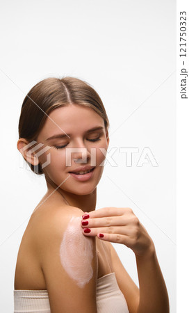 Smiling woman applying cream to her shoulder, symbolizing moisturizing and skincare routines against white studio background. Concept of natural beauty. 121750323