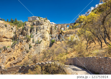 The Puente Nuevo bridge in El Tajo gorge in Ronda town in Spain. Ancient bridge spanning a dramatic gorge, with a village perched on the cliffs. A sunny day in a historical location. The Puente Nuevo bridge in El Tajo gorge in Ronda town in Spain. Ancient bridge spanning a dramatic gorge, with a village perched on the cliffs. A sunny day in a historical location. 121753914