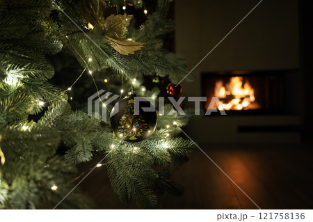 A close-up of a Christmas tree branch with a black ornament featuring a painted tree, illuminated by string lights, with a glowing fireplace in the background. A close-up of a Christmas tree branch with a black ornament featuring a painted tree, illuminated by string lights, with a glowing fireplace in the background. 121758136