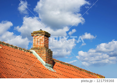A house with a red brick chimney on an asbestos slate roof outside with a blue sky background. Exterior building designed construction and architecture of a rooftop chute for fireplace smoke and heat 121759168