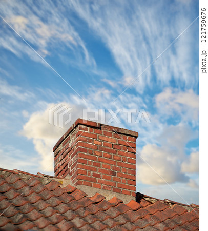 A brick chimney on the roof in the blue cloudy sky background. A closeup of the chimney on the rooftop. An orange bricked roof with chimney against the blue sky with copy space on a sunny day. 121759676