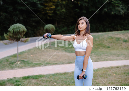 Young woman exercising with dumbbells in a park, promoting healthy lifestyle and fitness Young woman exercising with dumbbells in a park, promoting healthy lifestyle and fitness 121762159