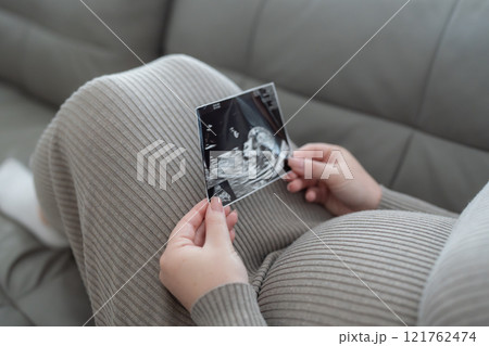 Gen Z pregnant woman holding ultrasound scan photo of a fetus sitting on a sofa. Concept of pregnancy, Maternity prenatal care 121762474