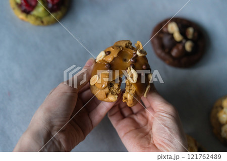 Woman breaks homemade american traditional chocolate chip cookie with peanut butter, sticky caramel and peanuts topping. Cookie broken in half.. 121762489