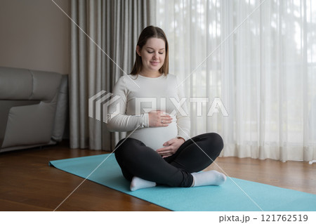 Caucasian young pregnant woman holding her belly while sitting on yoga mat at home. Workout, yoga and fitness at home, pregnancy concept. Pre-natal. Caucasian young pregnant woman holding her belly while sitting on yoga mat at home. Workout, yoga and fitness at home, pregnancy concept. Pre-natal. 121762519