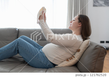 Pregnant woman holding baby onesie bodysuit sitting on a sofa at home. Preparing and planning baby clothes for new baby. Expectant mother. Pregnant woman holding baby onesie bodysuit sitting on a sofa at home. Preparing and planning baby clothes for new baby. Expectant mother. 121762520