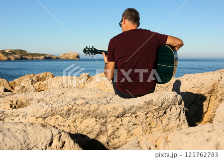 A man in a dark t-shirt playing acoustic guitar on a rocky area by the sea 121762783