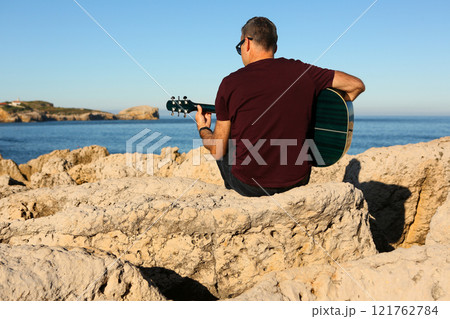 A man in a dark t-shirt playing acoustic guitar on a rocky area by the sea 121762784
