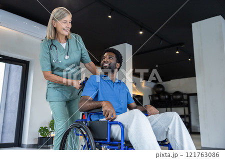 Mature female doctor carrying a wheeling-chair with a male patient 121763086