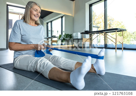 Mature woman using resistance band for stretching injured leg after trauma in rehabilitation center 121763108