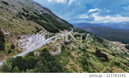 Winding mountain road leading through green valley under cloudy sky 121763242