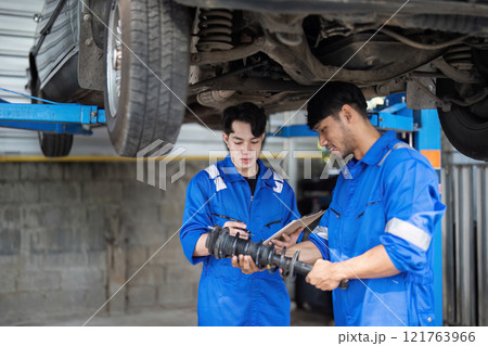 Young Man Repairing Car at Auto Repair Shop with Mechanic in Blue Uniforms, Inspecting Car Parts and Discussing Maintenance 121763966