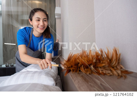 Smiling Maid Cleaning a Modern Room with Feather Duster in Hand, Wearing Blue Uniform and Apron Smiling Maid Cleaning a Modern Room with Feather Duster in Hand, Wearing Blue Uniform and Apron 121764102