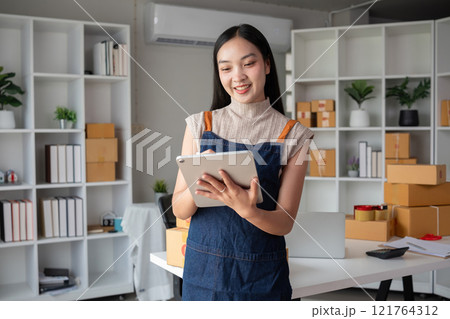 A small SME business owner receives product orders and writes shipping information on cardboard boxes in the home office to prepare for delivery. 121764312