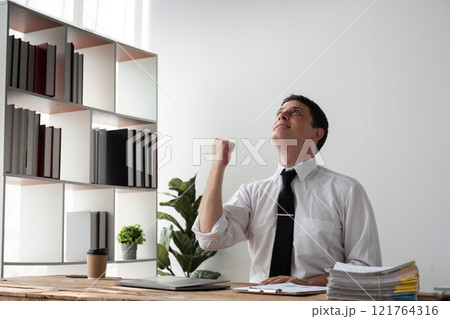 Young Businessman Celebrating Success in Modern Office with Joyful Expression and Raised Fist 121764316