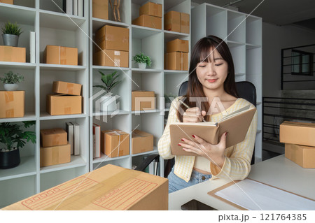 Young Woman Taking Online Sales Orders in Modern Home Office Surrounded by Shipping Boxes and Shelves 121764385