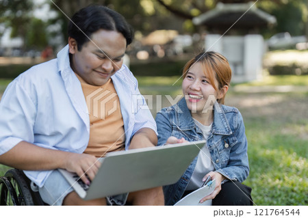 Young Man in Wheelchair Collaborating with Friend on Paperwork Outdoors in a Park Setting Young Man in Wheelchair Collaborating with Friend on Paperwork Outdoors in a Park Setting 121764544