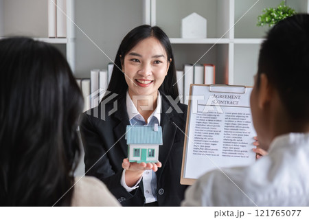 Happy Couple Signing Real Estate Sales Contract with Agent Holding Model House in Modern Office Setting 121765077