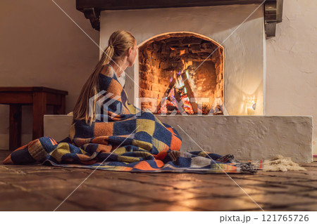 Woman by the fireplace during cozy winter evening. Warm and peaceful family moment indoors. Togetherness, love, and relaxation concept. A warm and inviting scene featuring a lit fireplace with glowing 121765726