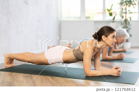 Three generations of women perform elbow plank Sampatasana in gym 121768612