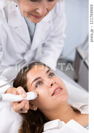 Elderly beautician doctor doing RF lifting procedure for flawless woman face laying in beauty salon. Hardware cosmetology. 121768669