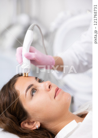 Unrecognizable beautician doctor doing RF lifting procedure for flawless woman face laying in beauty salon. Hardware cosmetology Unrecognizable beautician doctor doing RF lifting procedure for flawless woman face laying in beauty salon. Hardware cosmetology 121768731