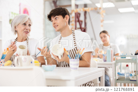 Cheerful elderly woman and guy painting ceramic mugs during pottery masterclass 121768891