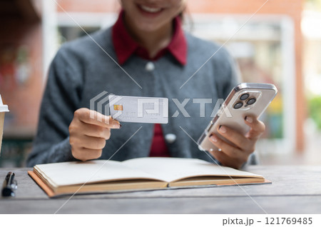 A cropped image of a woman holding a smartphone and a credit card while sitting at an outdoor table. A cropped image of a woman holding a smartphone and a credit card while sitting at an outdoor table. 121769485