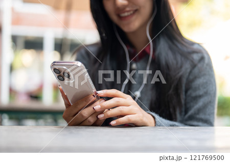 A woman enjoys listening to music on her earphones and using her smartphone while sitting outdoor. 121769500